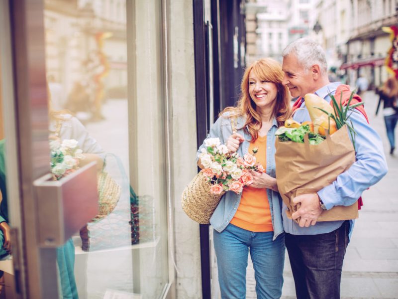 Mature couple outdoors on city street with grocery bags looking at shop display. They were in shop for breakfast. Enjoying in sunny weekend morning. Caucasian ethnicity.